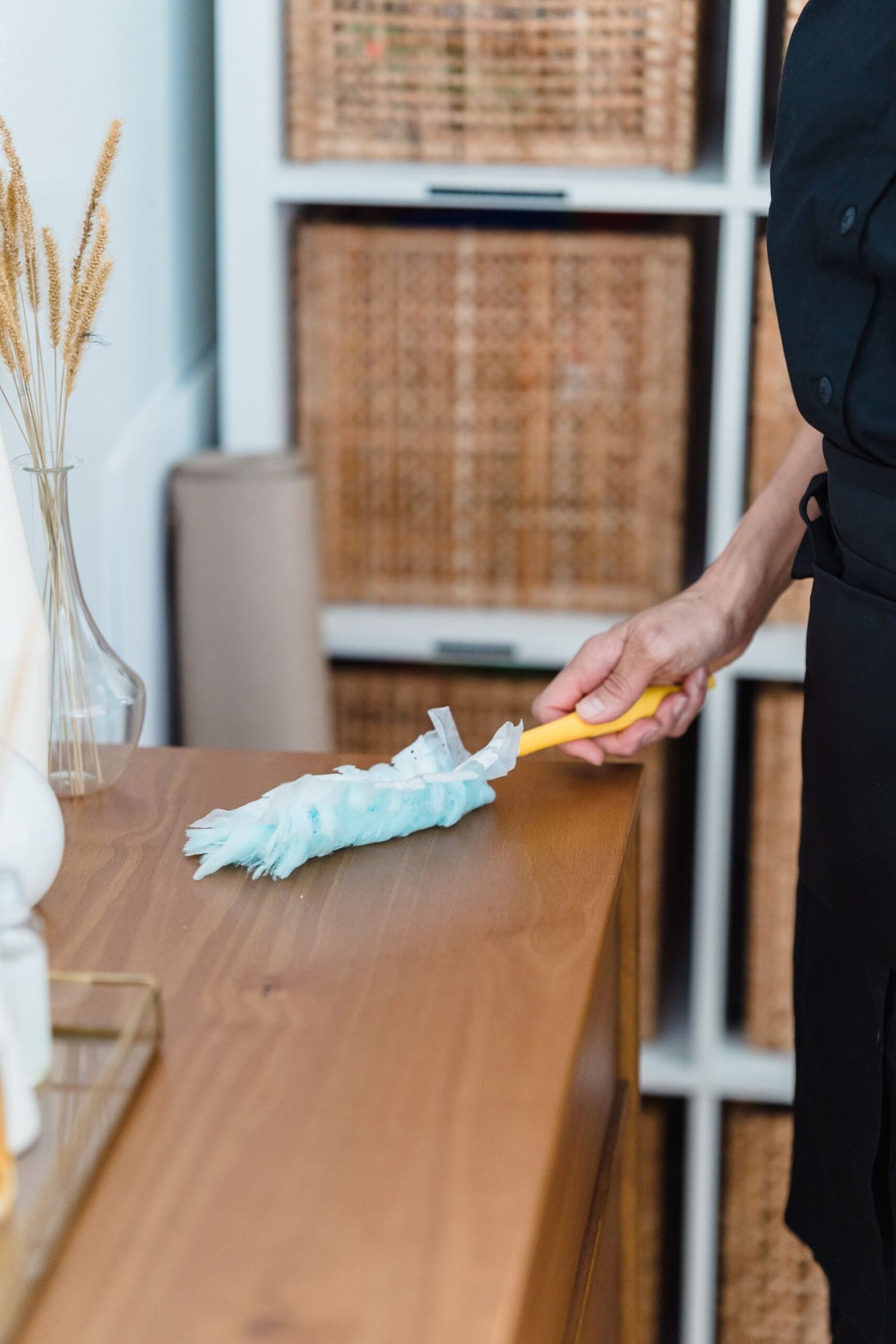 A woman in a black apron dusts a wooden dresser, highlighting hou
