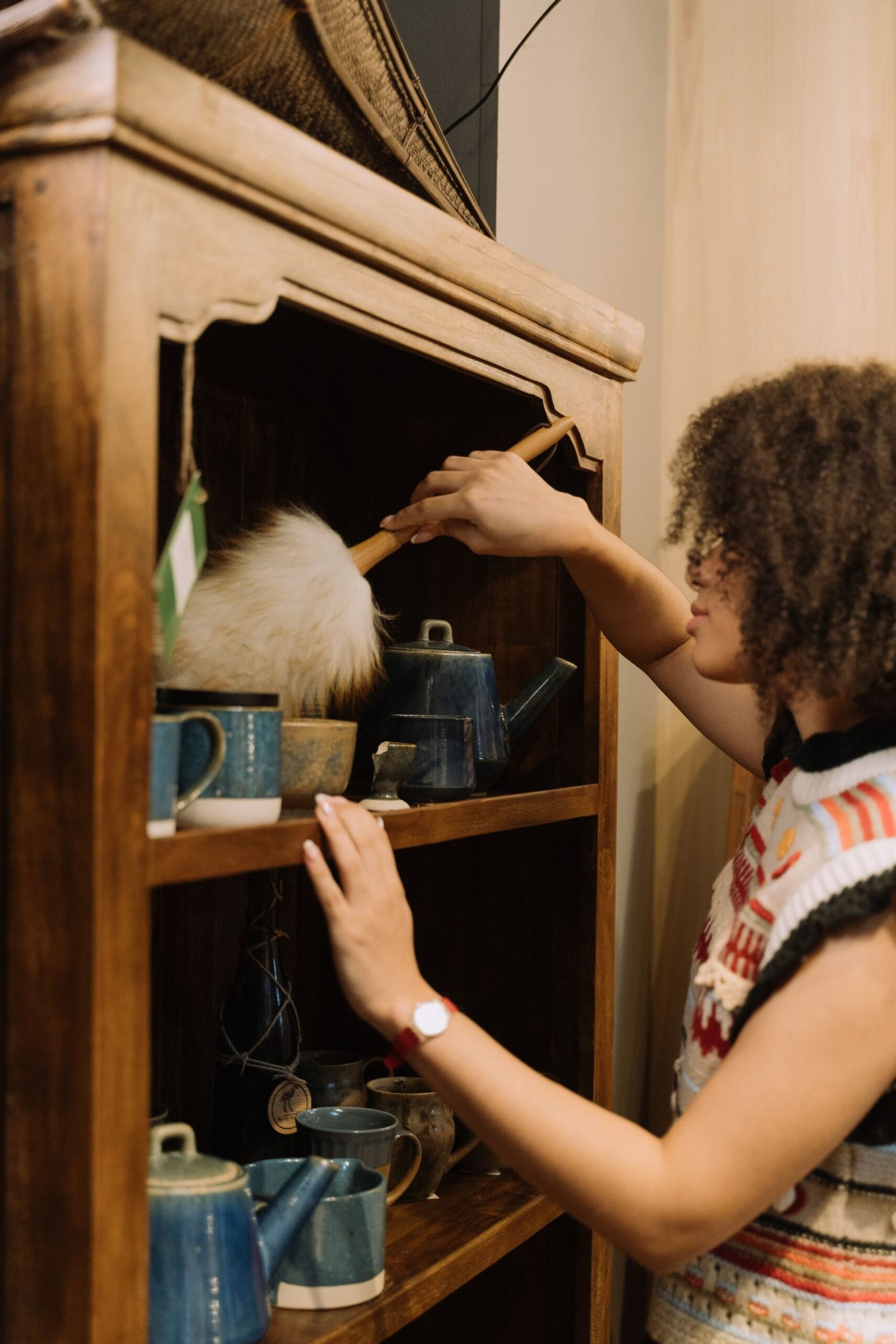Woman cleaning a wooden shelf filled with ceramic cups and teapot