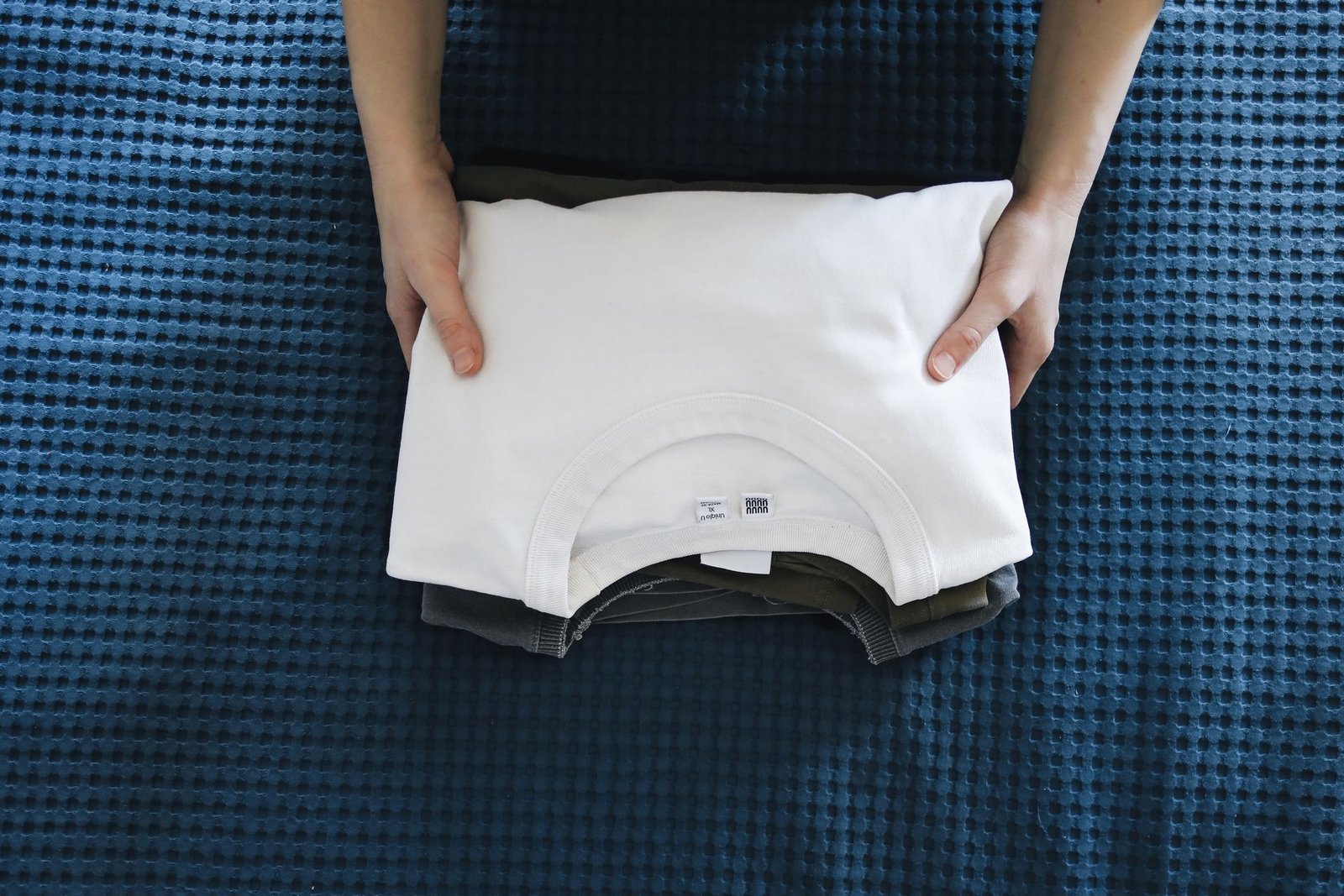 Hands folding a white shirt on a blue textured bedspread, focusin