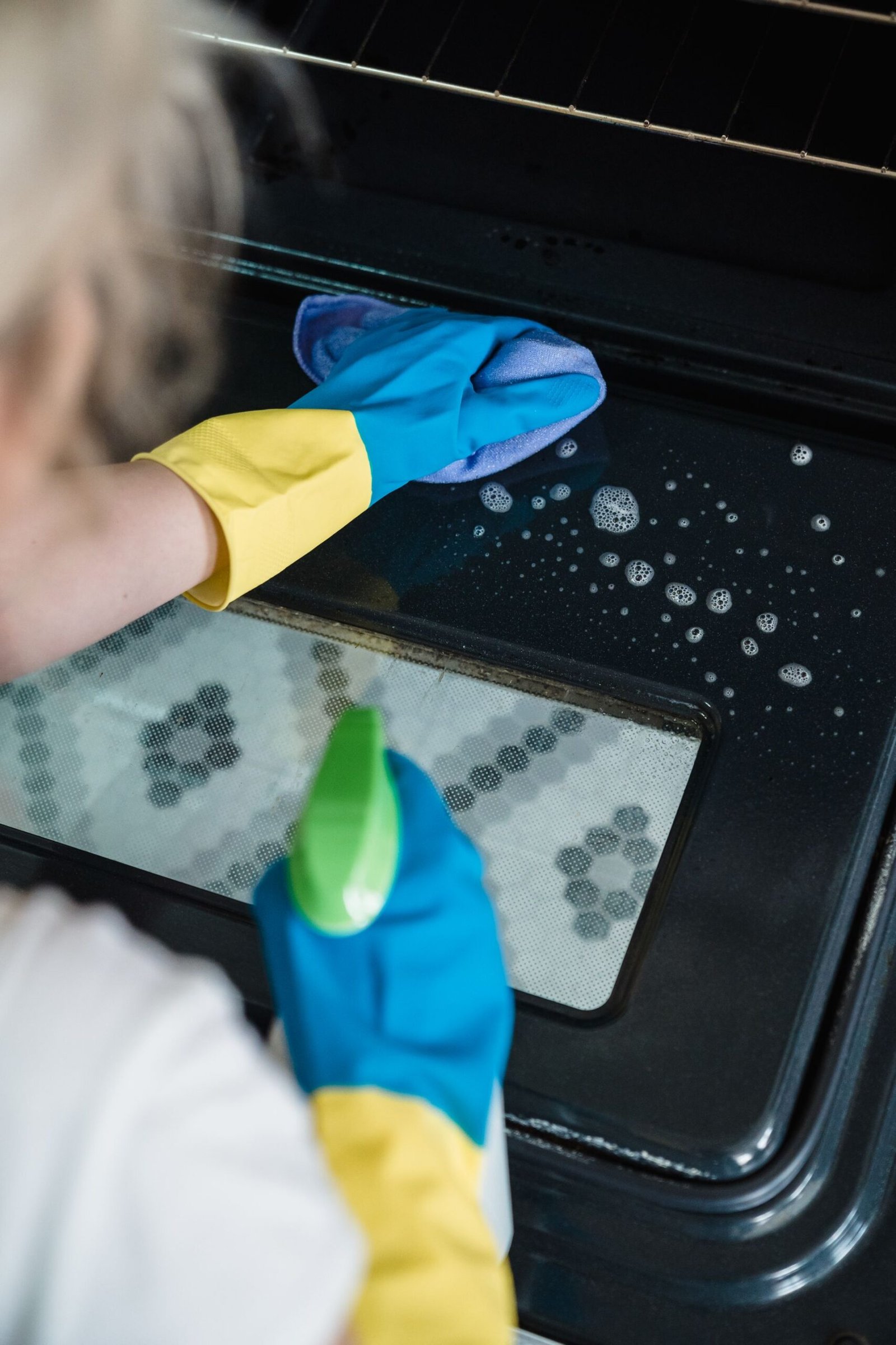 A person wearing gloves cleans an oven door using a spray and clo