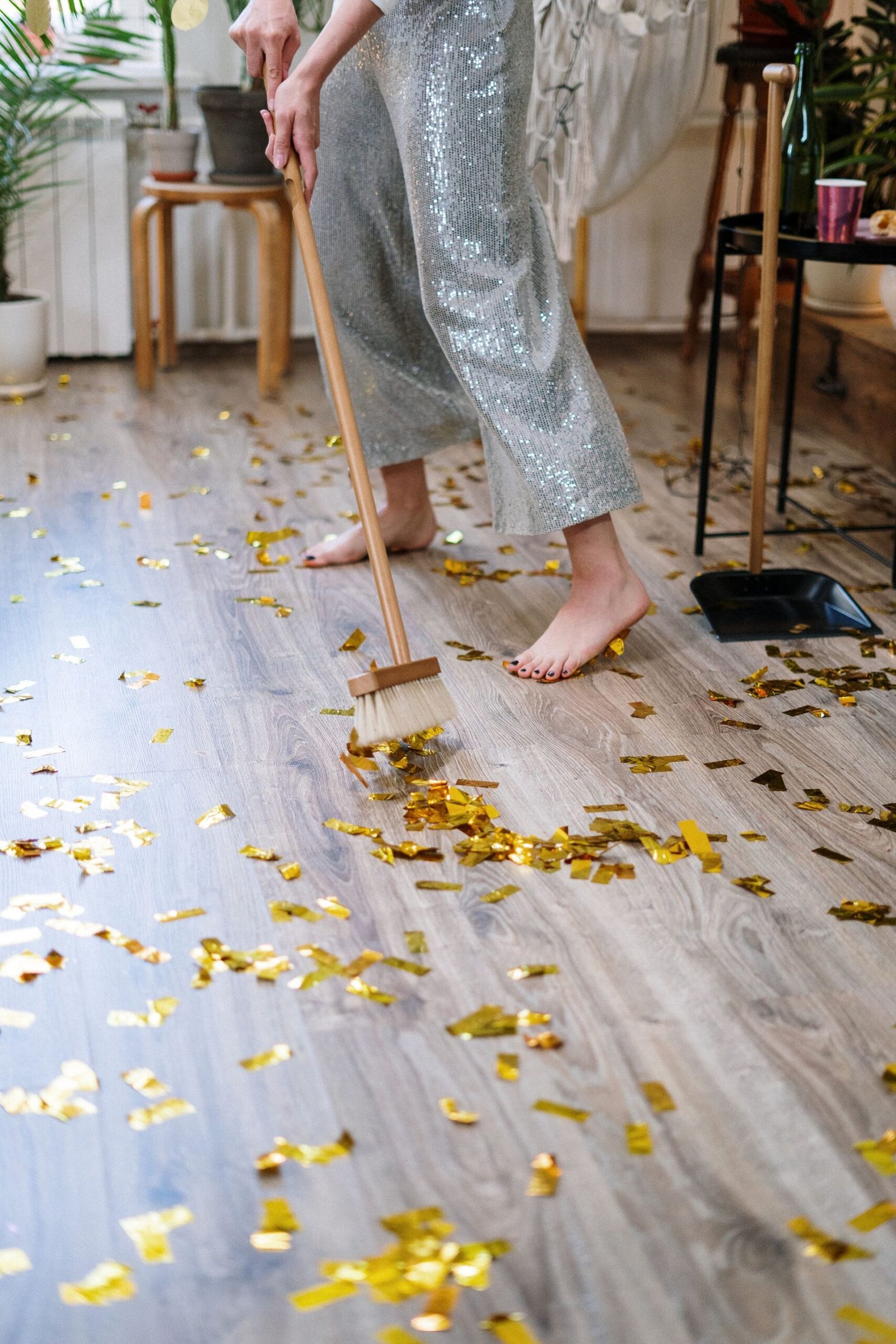 Person sweeping golden confetti off wooden floor after a party, b